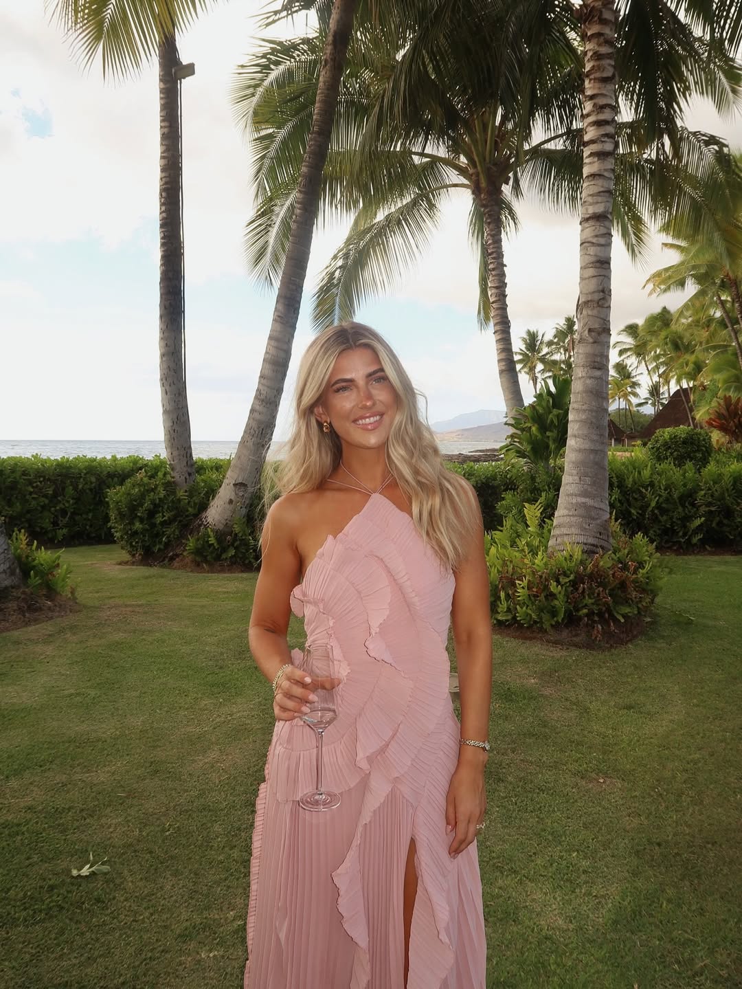 Woman smiling in the pink Millie Ruffle Midi Dress outdoors, with a pleated skirt and ruffled details.